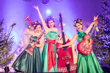 Zucchini Sistaz, Drei Frauen mit Posaune, Kontrabass, Gitarre auf einer Bühne. Alle Instrumente sind weihnachtslich eingestrickt. Im Hintergrund Weihnachtsbäume und Sterne. Die Frauen winken in die Kamera.